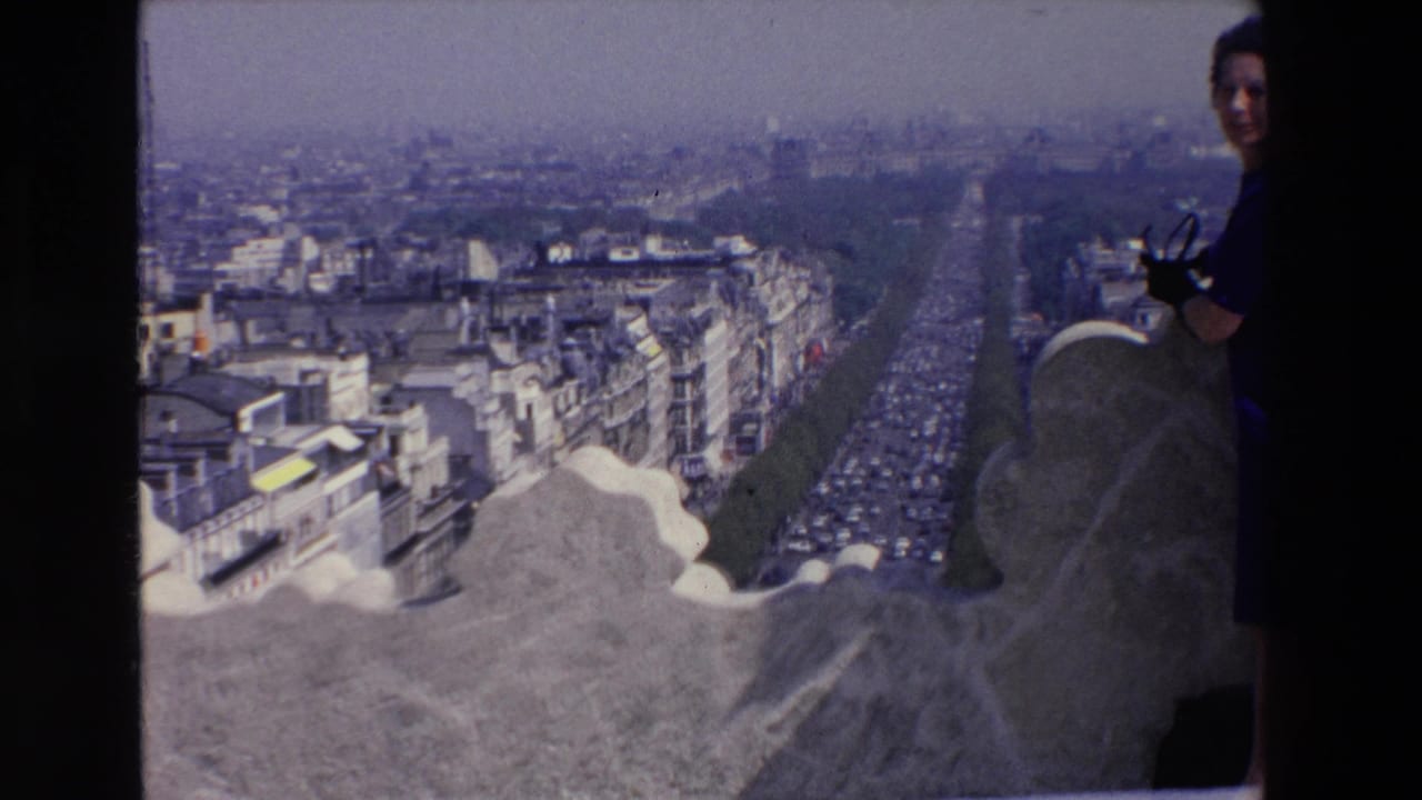 In 1969 France home-movie footage, a woman in a blue dress stands partly cut off beside a stone overlook with a broad city view behind her.
