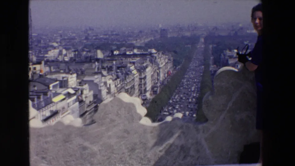 In 1969 France home-movie footage, a woman in a blue dress stands partly cut off beside a stone overlook with a broad city view behind her.