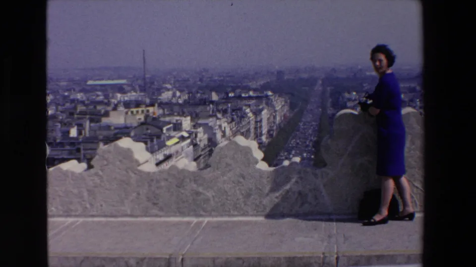A woman in a blue dress stands near a decorative stone wall at a high French city viewpoint, with a long traffic-filled avenue receding into haze.