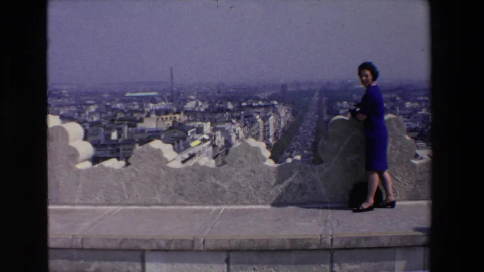 The woman turns toward the movie camera while holding a camera of her own on a 1969 French observation terrace above dense city streets.