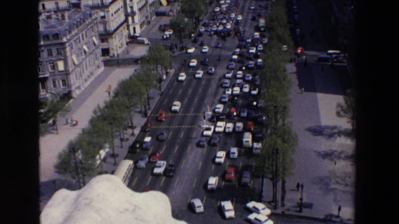 1969 8mm frame from Loudes, France, showing an elevated view over a broad boulevard with dense car traffic, sidewalks, street trees, and pale multi-story buildings.