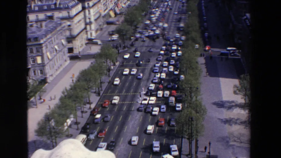 High-angle 1969 home-movie view in Loudes, France, with compact cars clustered in traffic lanes, rows of trees, sidewalks, and older urban facades.