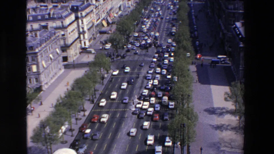 Elevated 8mm frame of a French city boulevard in 1969, with traffic flowing and queuing between sidewalks, street trees, and multi-story stone buildings.