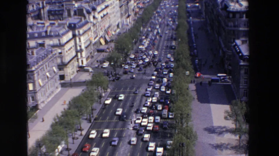 Wide overhead 1969 8mm view from Loudes, France, showing a busy boulevard packed with cars, flanked by sidewalks, trees, and urban stone architecture.
