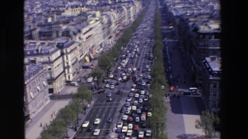 Elevated view of late-1960s city traffic in France, with rows of cars moving along a tree-lined boulevard beside ornate urban buildings.