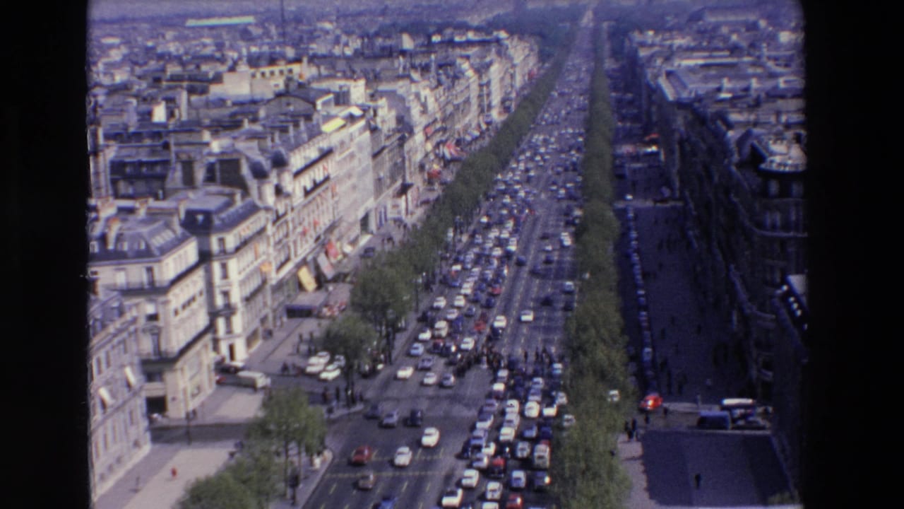 High-angle 8mm view of a congested French boulevard in the 1969 clip metadata, with period cars, tree rows, sidewalks, and stone urban facades.