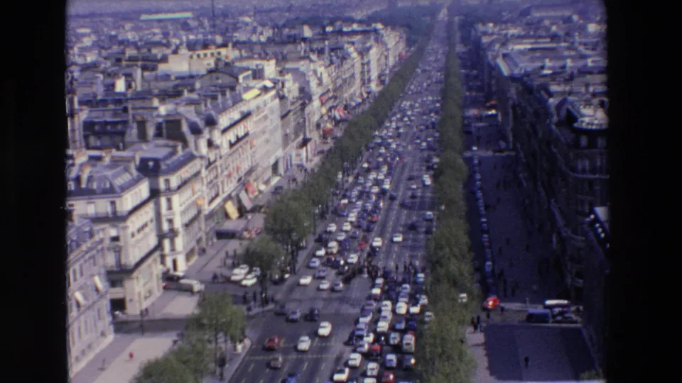 High-angle 8mm view of a congested French boulevard in the 1969 clip metadata, with period cars, tree rows, sidewalks, and stone urban facades.