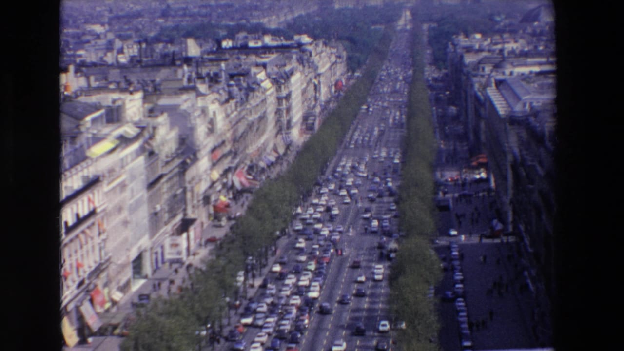 Elevated view of a long Parisian-style boulevard with slow traffic, tree rows, pedestrians at sidewalk scale, and late-1960s urban architecture visible in the supplied clip context.
