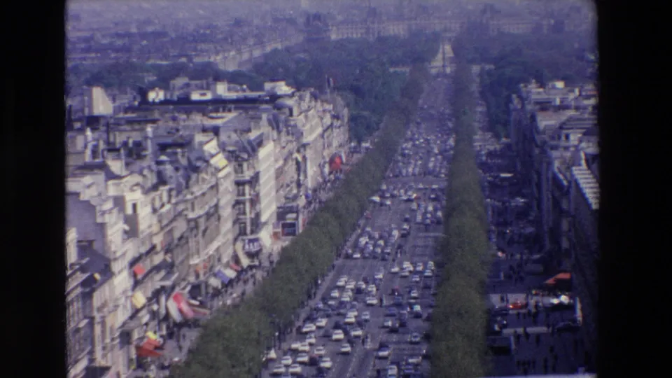 Soft 8mm frame of a congested French city boulevard, with pale masonry buildings, awnings, tree-lined lanes, and compact cars stretching into the distance.