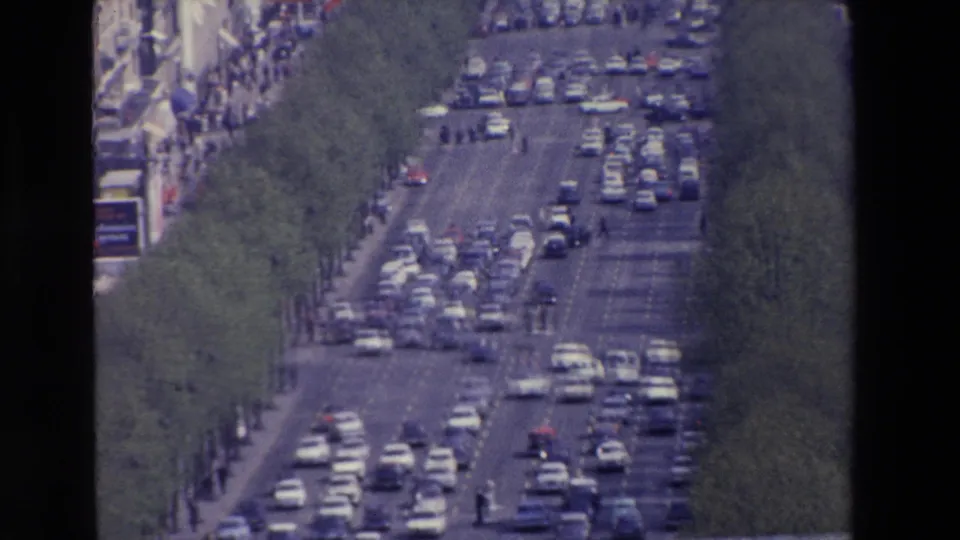 Closer 8mm view down the same congested boulevard, emphasizing rows of cars, green trees, lane markings, and small pedestrian figures along the street edges.