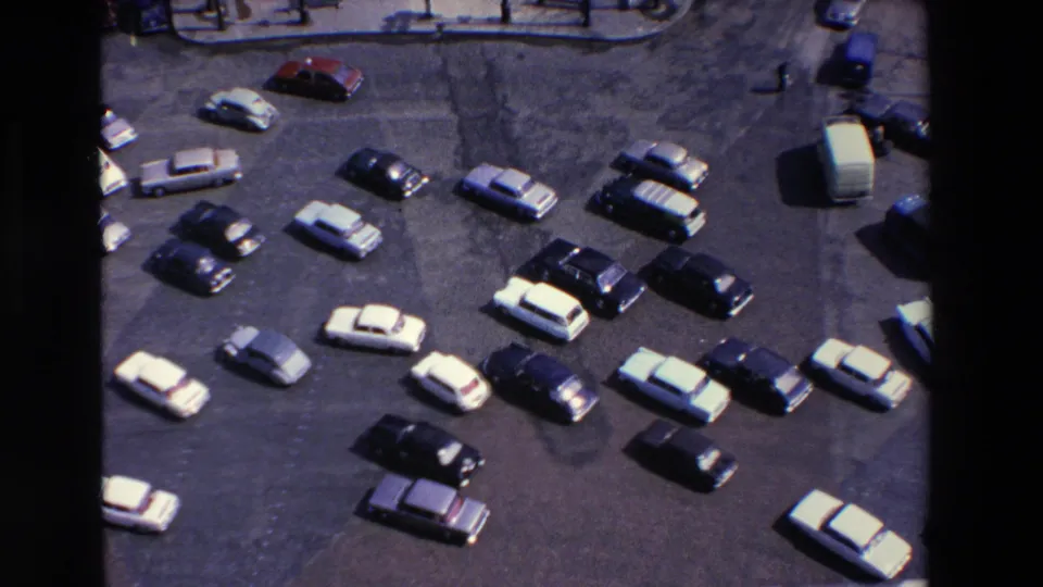 Overhead home-movie frame from the 1969 Loudes, France clip showing rows of parked cars, a pale van, and a distant walkway along the top of the lot.