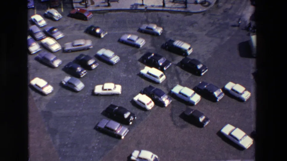 Elevated 8mm frame showing clustered 1960s-era cars in a paved parking area, with soft color and black film borders visible in the scan.