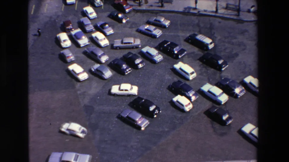 High-angle 1969 Loudes, France frame with a busy parking lot, varied sedans and wagons, a small white car near the lower edge, and a distant pedestrian near the top.