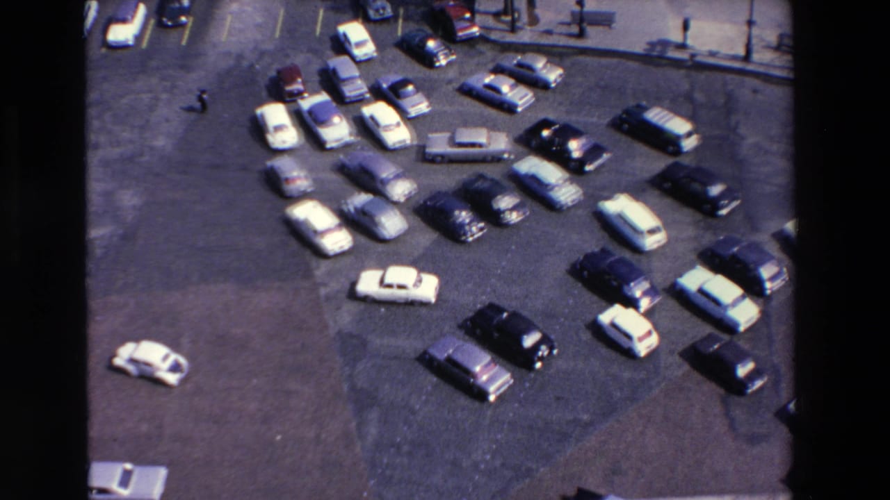 Elevated 1969 8mm view in Loudes, France, showing light and dark vintage cars scattered across a paved parking lot near a curb with lamps and benches.