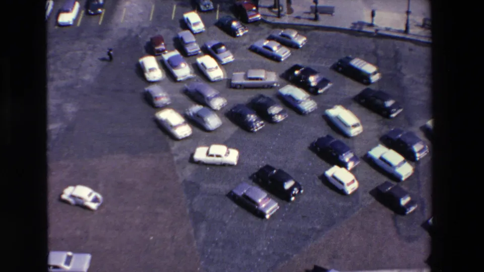 Elevated 1969 8mm view in Loudes, France, showing light and dark vintage cars scattered across a paved parking lot near a curb with lamps and benches.