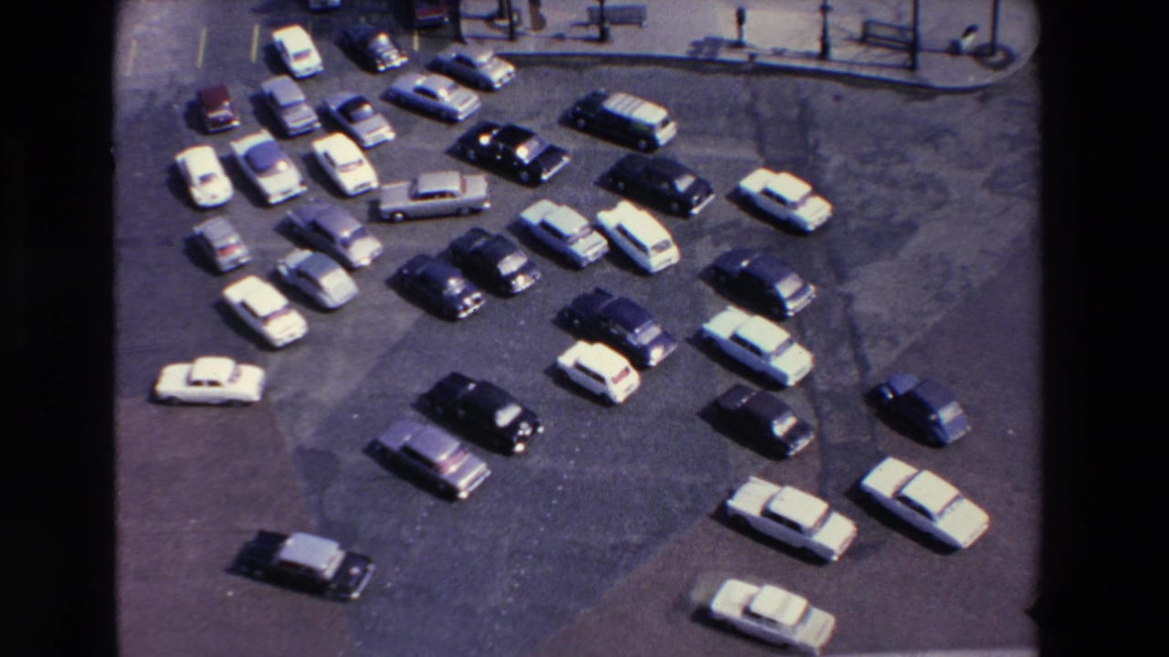 High-angle 1969 home-movie frame from Loudes, France, with sedans and station wagons parked in an open lot beside a paved street edge.