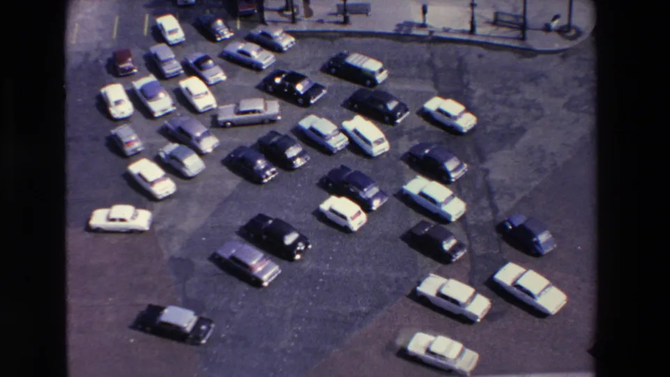 High-angle 1969 home-movie frame from Loudes, France, with sedans and station wagons parked in an open lot beside a paved street edge.