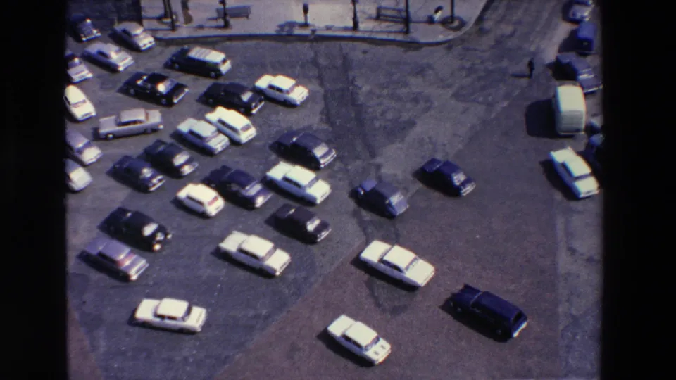 Elevated 1969 view in Loudes, France, showing a busy parking lot with multiple vintage cars, open pavement, curbside benches, and part of a bus at the right edge.
