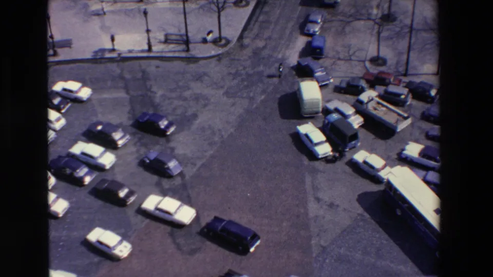 Late-1960s 8mm frame from Loudes, France, with cars spread across a paved lot, a large bus entering the lower right, and small pedestrians near the curbside trees and benches.