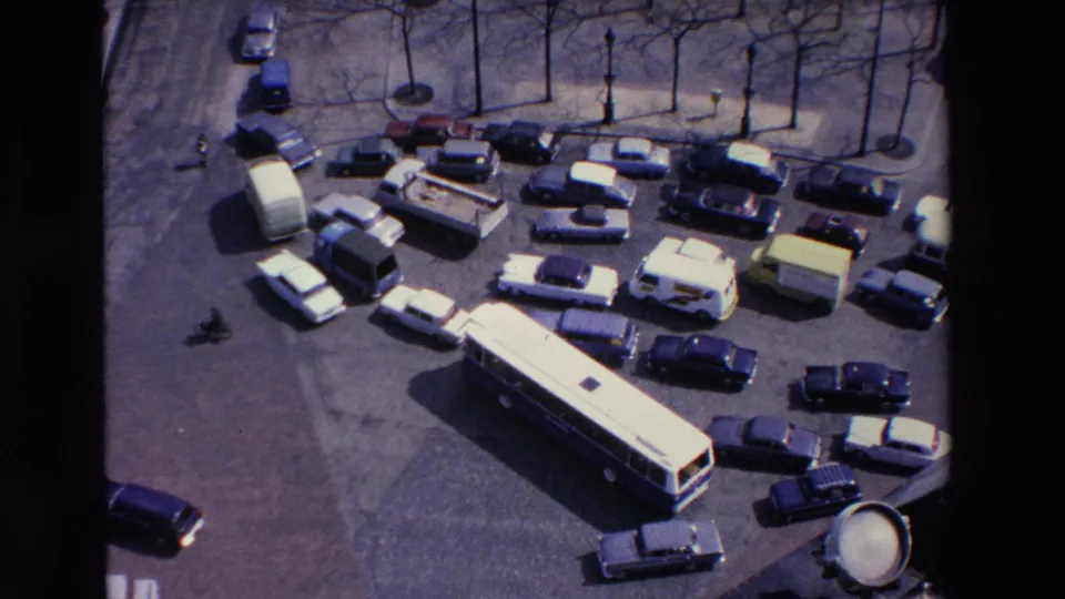 High-angle frame from the supplied 1969 France clip showing a long bus, compact cars, vans, and a small motorcyclist within a congested paved street area.