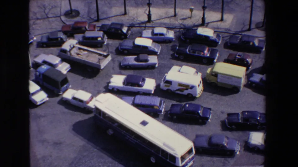 High-angle 8mm frame of late-1960s traffic in France, with a bus, compact cars, vans, and a small truck packed into a city street scene.