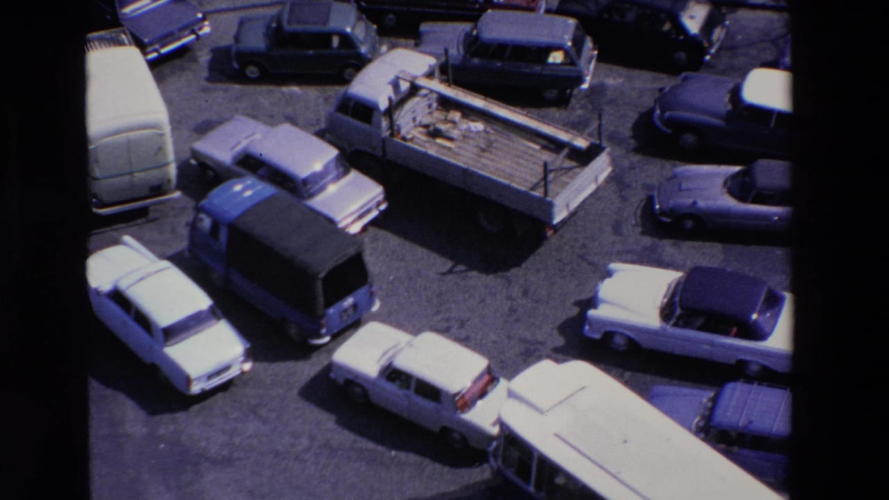 Elevated 1969 Loudes, France 8mm frame showing sedans, vans, and a flatbed truck clustered around a sunlit paved intersection.