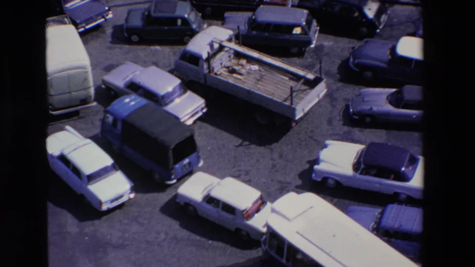 Elevated 1969 Loudes, France 8mm frame showing sedans, vans, and a flatbed truck clustered around a sunlit paved intersection.