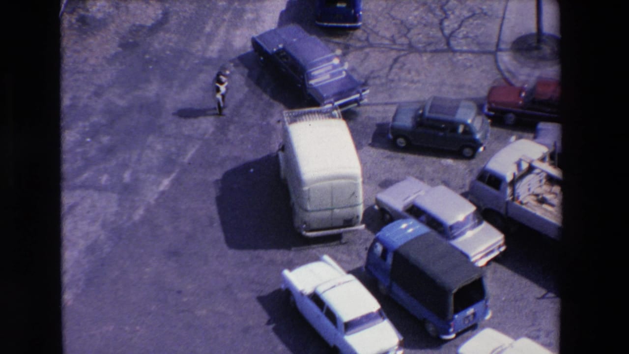 High-angle 1969 Loudes, France frame with a white van, several sedans, a flatbed truck, and a pedestrian visible near the open roadway.