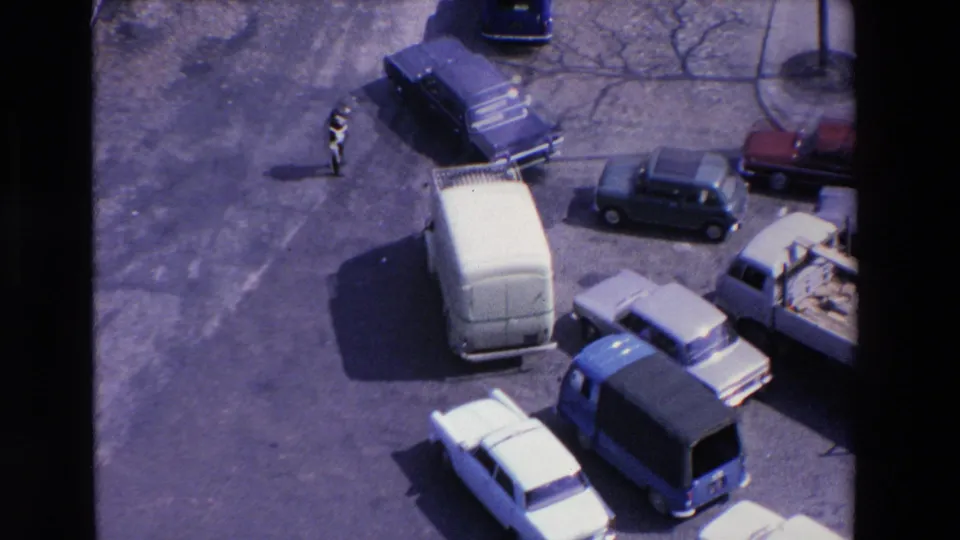 High-angle 1969 Loudes, France frame with a white van, several sedans, a flatbed truck, and a pedestrian visible near the open roadway.