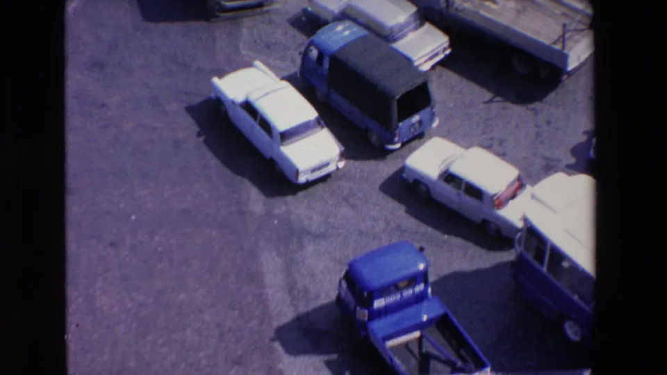 Elevated 1969 Loudes, France frame showing a white car, blue van, station wagon, and service vehicles moving through a busy paved traffic area.