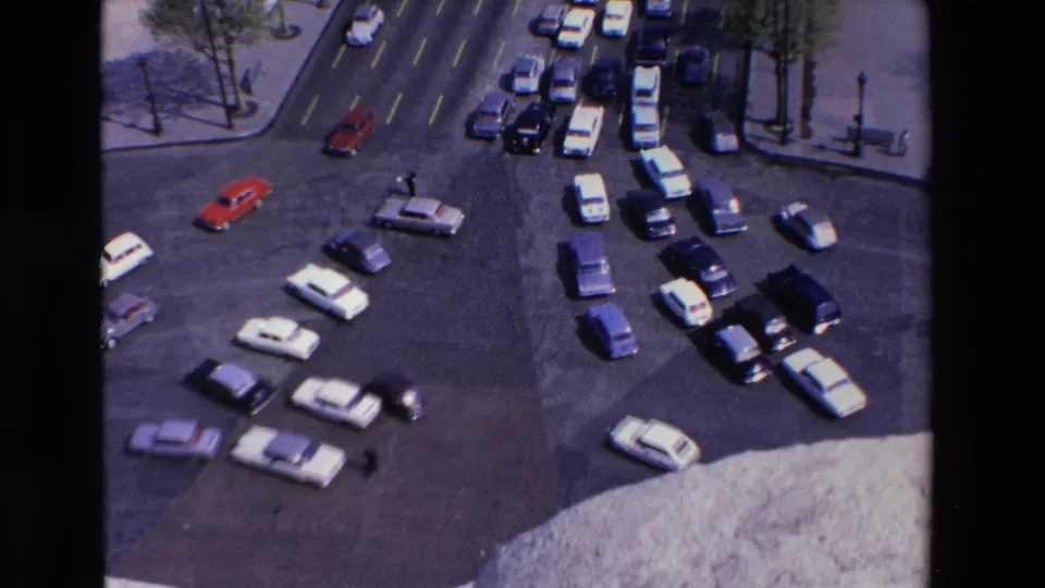Wide elevated 1969 Loudes, France frame showing many cars moving through a broad intersection with lane markings, roadside trees, and small pedestrian figures.