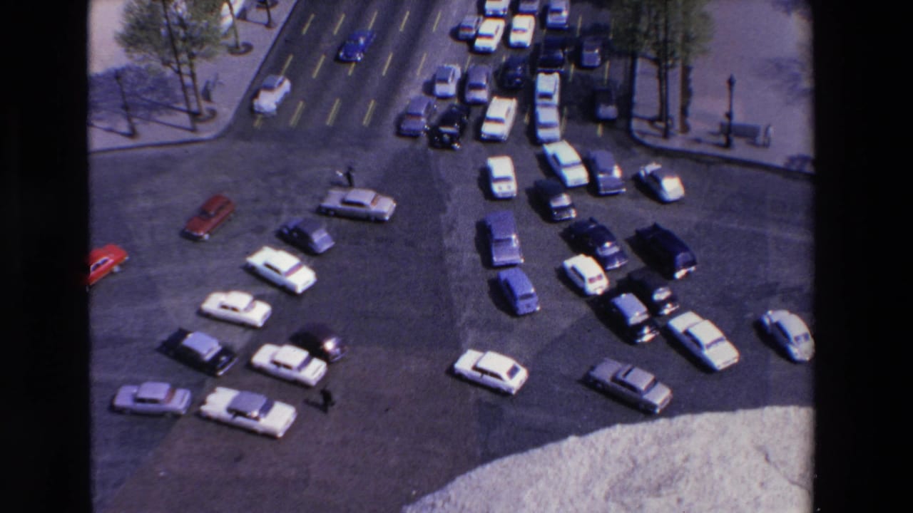 Elevated 1969 France 8mm view of a busy roundabout where white, red, and dark cars circulate near tree-lined sidewalks and a few pedestrians.