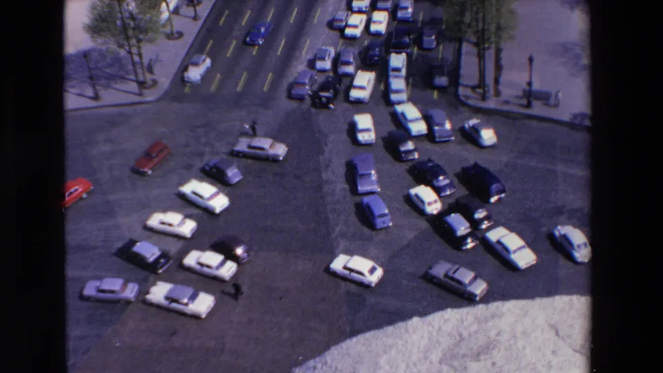 Elevated 1969 France 8mm view of a busy roundabout where white, red, and dark cars circulate near tree-lined sidewalks and a few pedestrians.