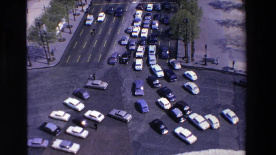 High-angle 1969 8mm frame of cars entering, exiting, and queuing around a French roundabout, with pedestrians small in the scene and tree-lined pavement visible.