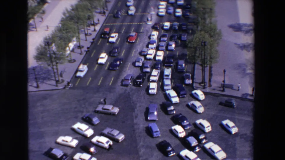 Elevated 8mm view of a congested roundabout approach in the France location identified by metadata, with rows of late-1960s cars, sidewalks, trees, and small pedestrians.