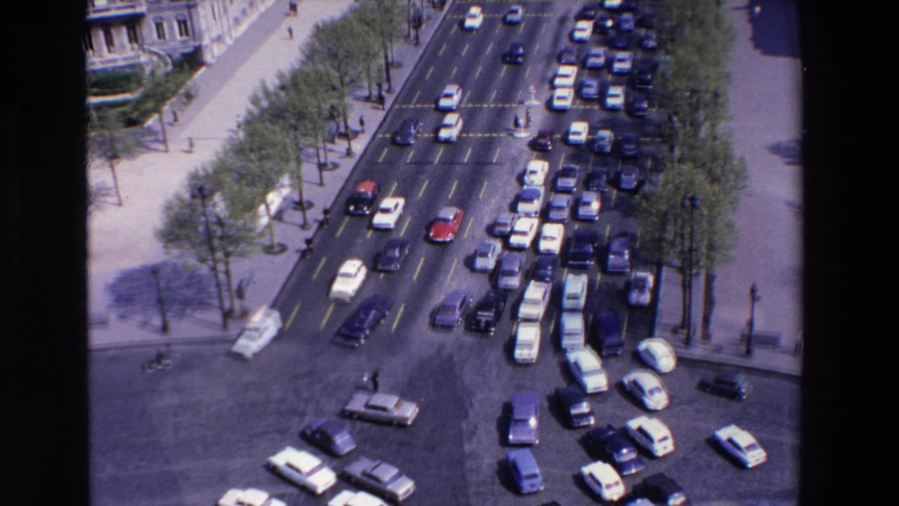High-angle 1969 France street view showing dense car traffic, pedestrians, tree-lined sidewalks, and ornate building facades at a broad urban intersection.