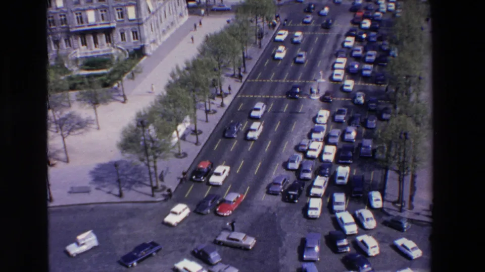 Elevated 1969 France view of cars queuing and turning through a busy boulevard intersection beside trees, sidewalks, and stone building fronts.