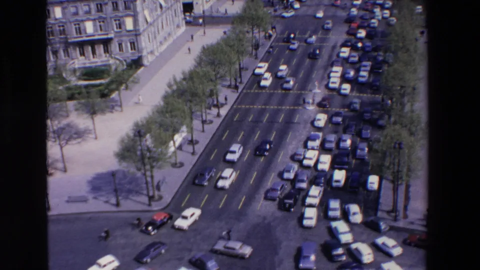 High-angle 1969 France frame showing cars spread across a broad intersection, with pedestrians, trees, sidewalks, and a large building edge visible around the roadway.