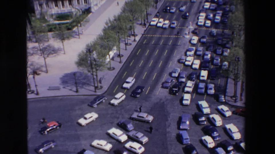 Elevated 1969 France view of multi-lane traffic, including white and dark cars, a red car, lane markings, trees, and pedestrians near a broad intersection.