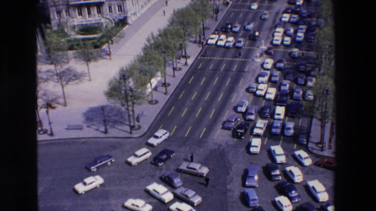 Elevated 1969 France home-movie view of a broad city intersection with moving cars, parked traffic, sidewalks, trees, and stone buildings.