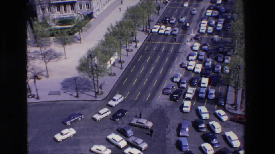 Elevated 1969 France home-movie view of a broad city intersection with moving cars, parked traffic, sidewalks, trees, and stone buildings.