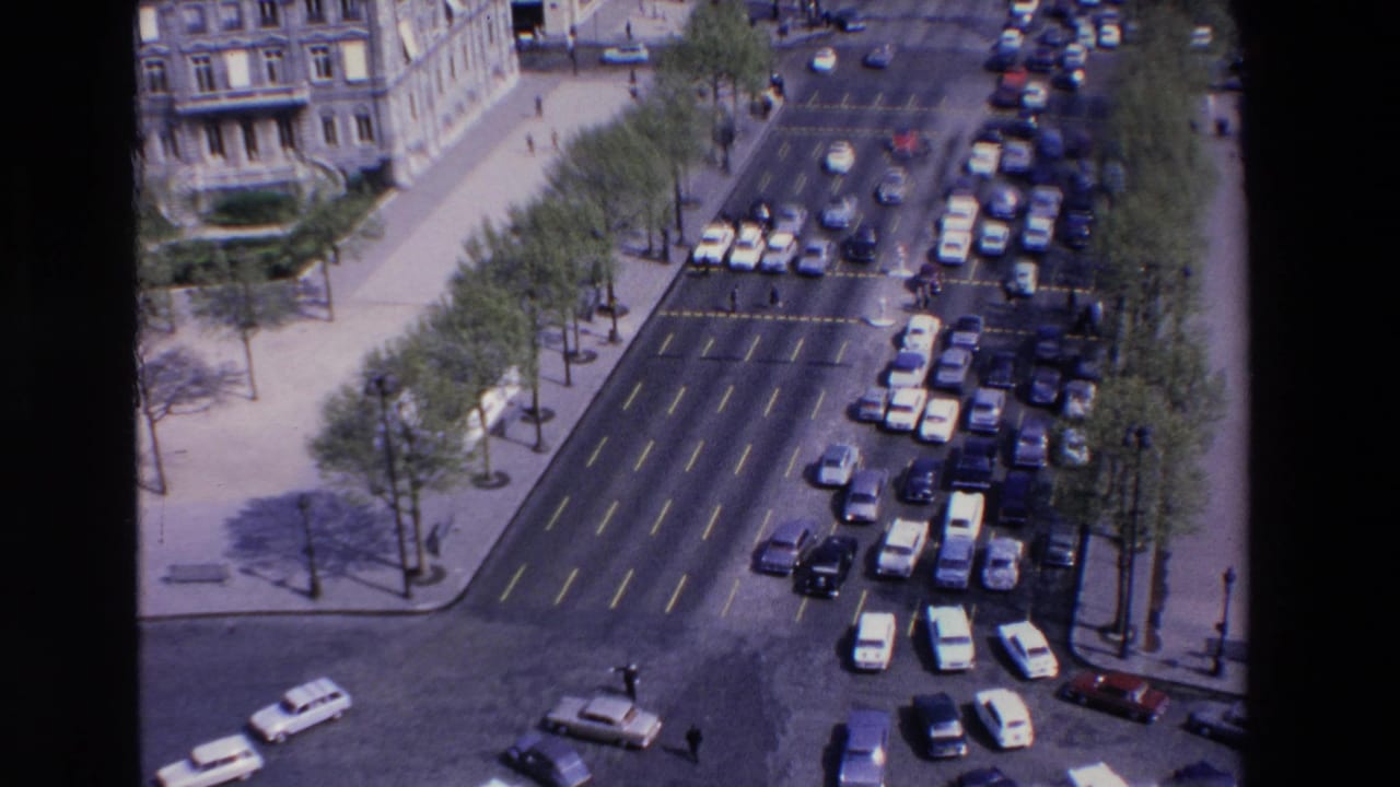 Elevated home-movie frame from the stated 1969 France clip showing dense car traffic, wide lanes, sidewalks, trees, and pale stone architecture.