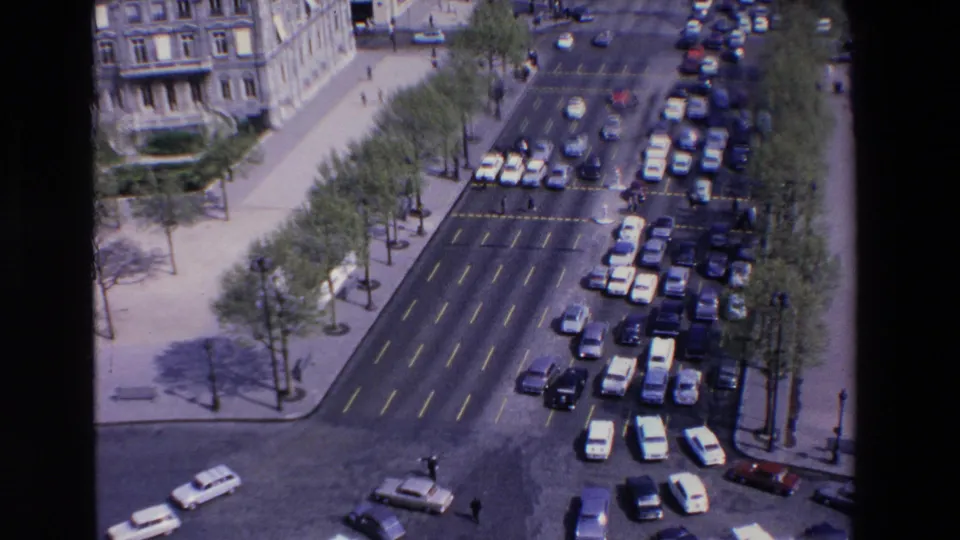 Elevated home-movie frame from the stated 1969 France clip showing dense car traffic, wide lanes, sidewalks, trees, and pale stone architecture.