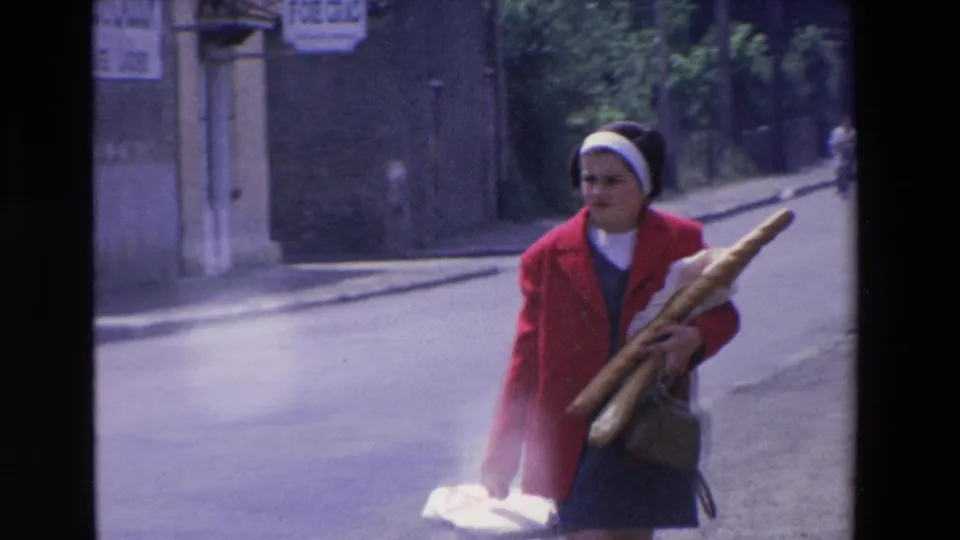 A woman in a red jacket and white headband walks along a French street in the stated 1969 clip while carrying long loaves of bread under one arm.