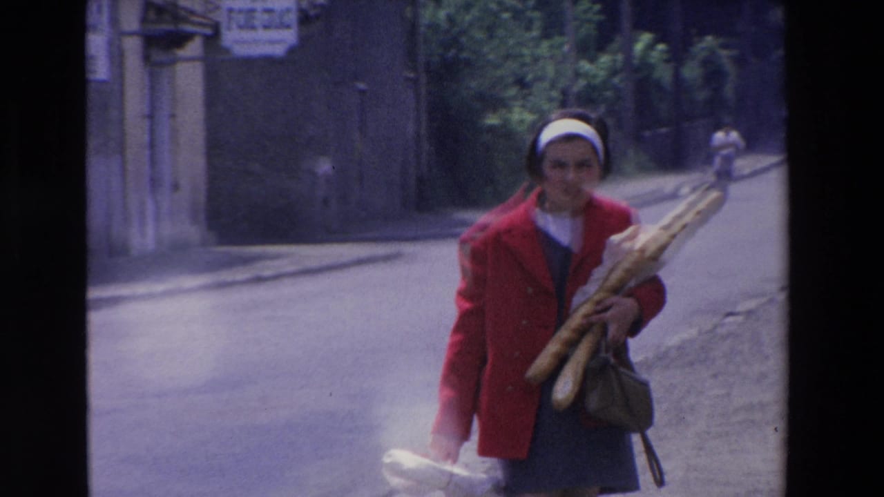 In Loudes, France, a woman in a red coat and white headband walks along a quiet street carrying several long baguettes in 1969 home-movie footage.