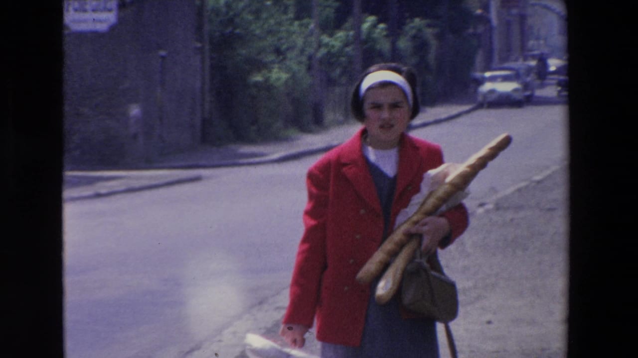 A woman in a red coat holds several baguettes and a handbag while walking toward the camera on a quiet Loudes street, consistent with the supplied 1969 metadata.