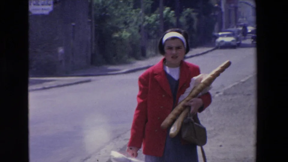 A woman in a red coat holds several baguettes and a handbag while walking toward the camera on a quiet Loudes street, consistent with the supplied 1969 metadata.