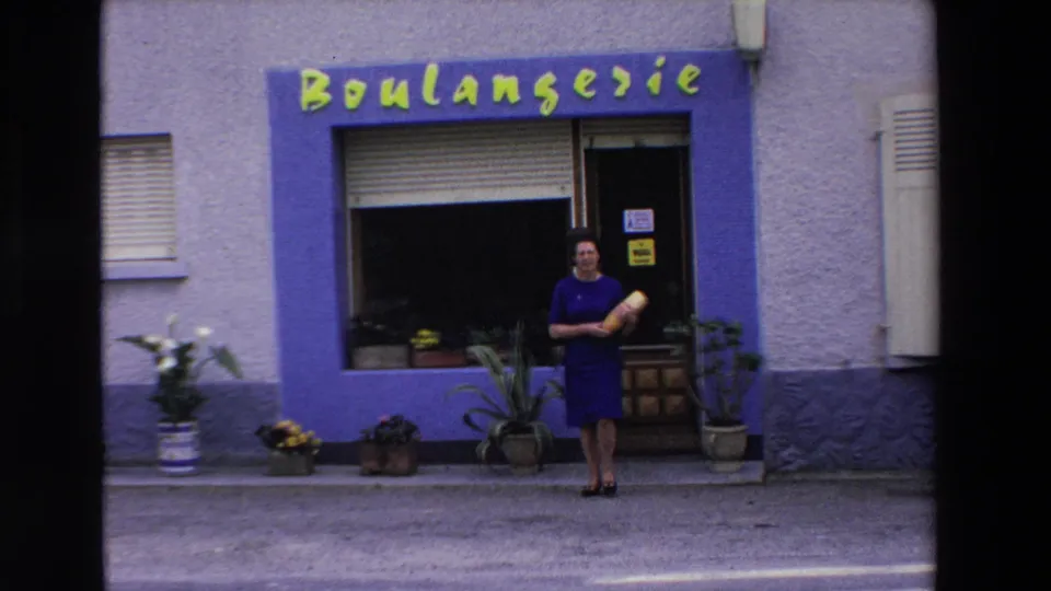 A woman holding a baguette stands in front of a purple-trimmed boulangerie with yellow signage, potted plants, and a recessed doorway in Loudes, France.