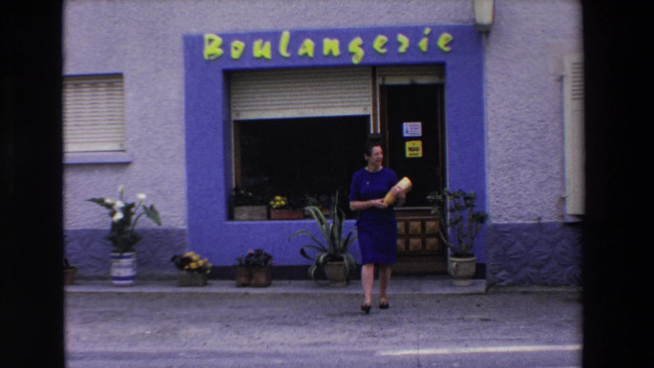 Woman in a purple dress stands outside a boulangerie holding a baguette.