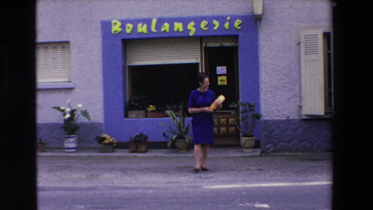 A 1969 Loudes, France home-movie frame shows a woman carrying a baguette near an open boulangerie doorway with storefront lettering and plants along the sidewalk.